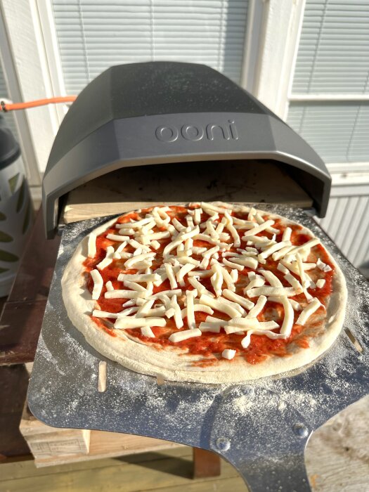 Ooni ugn med oostad pizza på pizzaspad, redo för gräddning. Tomatsås och ost på degen.