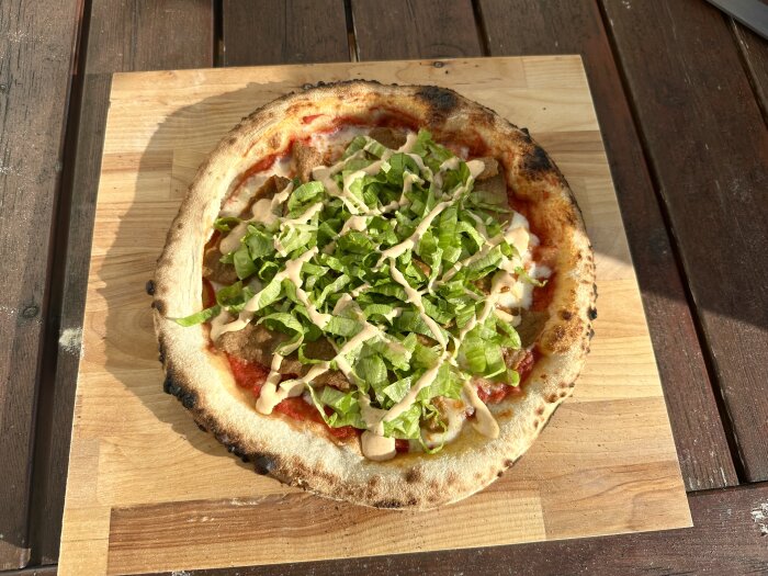 Nygräddad pizza med sallad ovanpå, serveras på träbräda, utomhus i solljus.