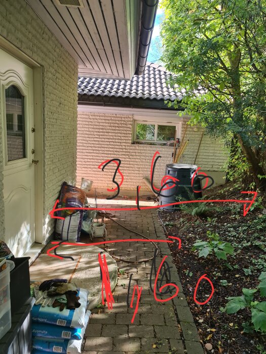 Trädgårdsgång med byggmaterial, mätningar markerade med rött, växtlighet, träd, och en husfasad.
