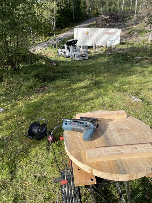 Arbetsplats utomhus: verktyg, trä, bil, skog, container med text "RETURKÄRL", arbetsbord, hörselskydd.