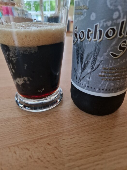 Mörkt öl i glas bredvid en del av ölflaska på träbord. Ljus skumkrona syns.