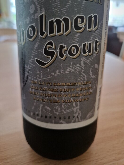 Ölflaska, "Holmen Stout", beskrivning på svenska, mörk bakgrund, delvis skarp fokusering, inomhusmiljö.