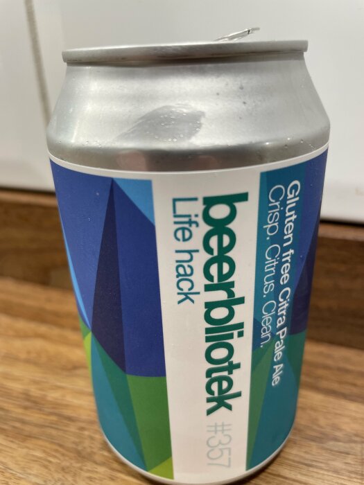 Aluminiumburk för dryck, färgglad etikett med text, står på träyta, öppnad lock, skadad öppning, öl eller läsk.