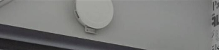 En vit parabolantenn monterad på en vägg under ett grått tak, otydliga ord till höger.