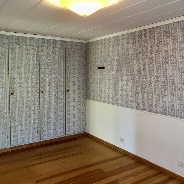 Ett tomt rum med trägolv, mönstrad tapet, garderober och en taklampa.
