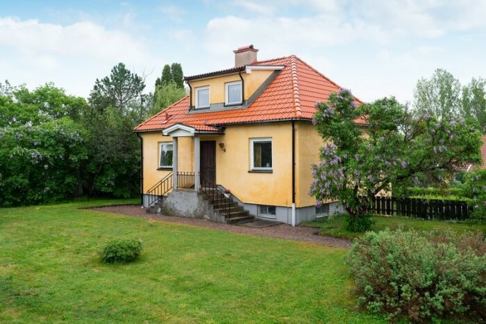 Gult hus, röda takpannor, grönt gräs, buskar, blommande träd, molnig himmel.