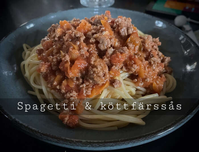En skål med spaghetti och köttfärssås på ett bord, hemmamiljö, text på bilden.