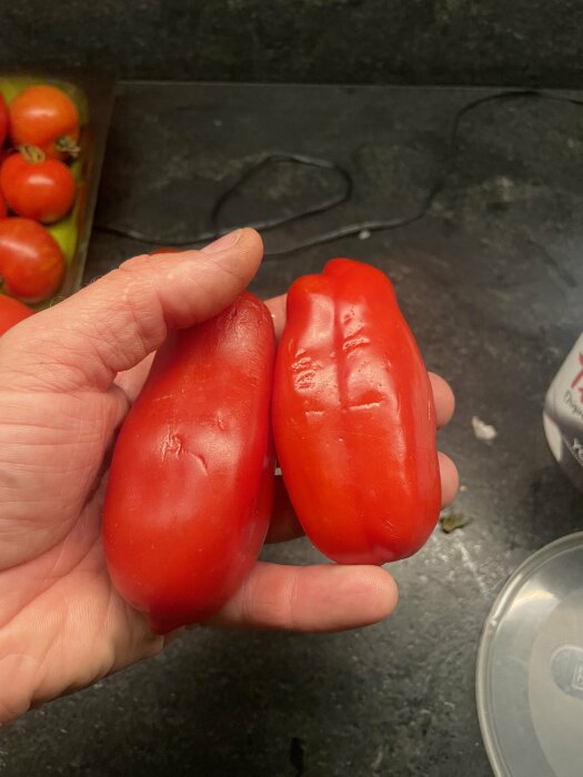 Hand som håller två röda, mogna San Marzano-tomater ovanför mörkt köksbänk med fler tomater i bakgrunden.