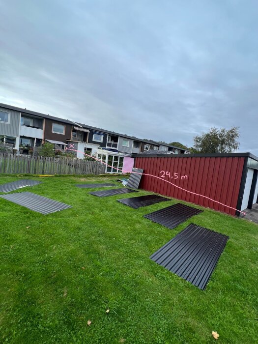 En röd container och svarta paneler på gräsmatta framför tvåvåningshus under molnig himmel.