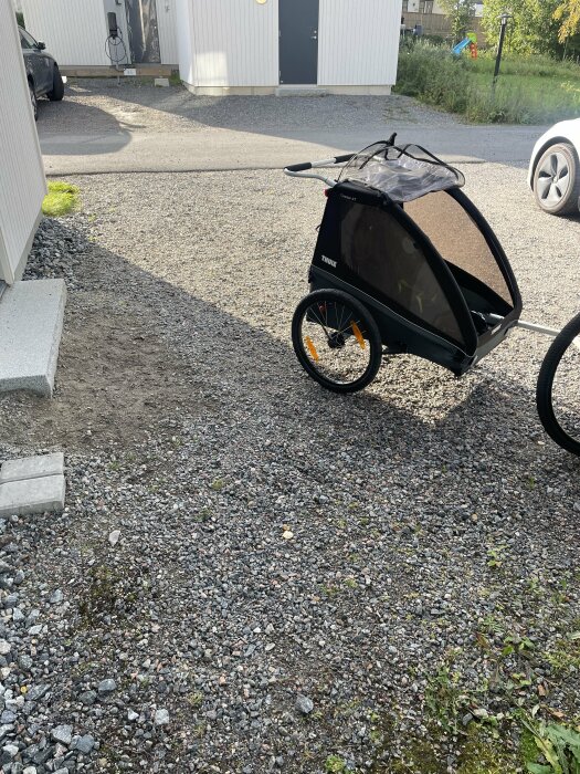 Cykelvagn på grusgång bredvid trappa, byggnader och parkerade bilar i bostadsområde under dagtid.