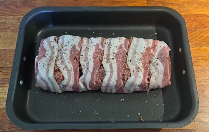 Rå baconlindad köttfärs i ugnsform, kryddad, redo för tillagning.