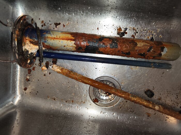 Rostig avloppsstoppning i ståldiskho med förfallna rör och smuts samlad runtomkring. Behöver reparation eller utbyte.