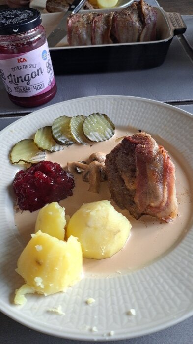 Ett måltidsfat med köttinlindad filé, potatis, lingonsylt, inlagda gurkor och gräddsås. Bakgrunden visar ytterligare mat.