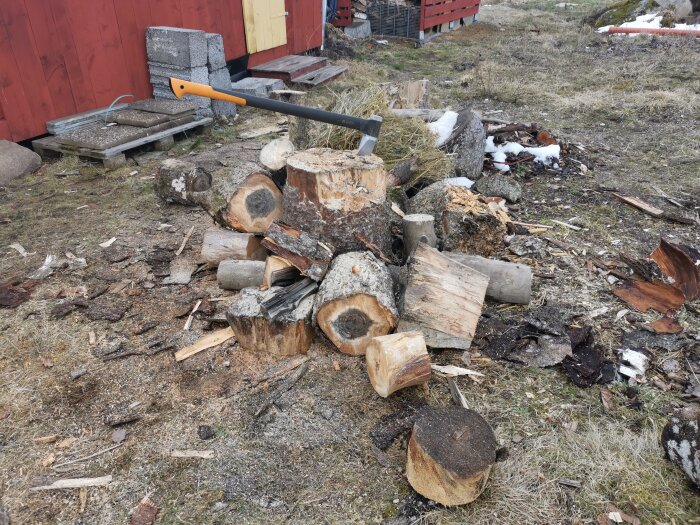 Vedhögar, kluven och okluven ved, yxa, utomhus, avfall, röd byggnad, gräsmatta, träklyvning.