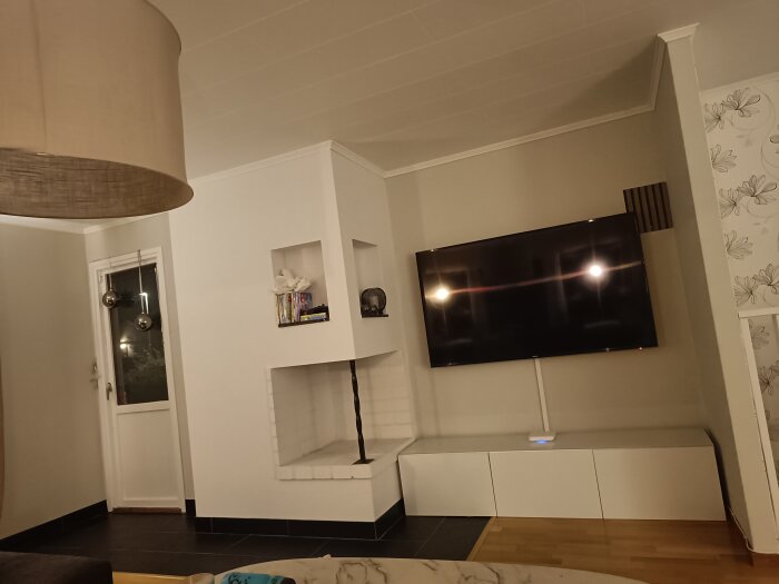 Modernt vardagsrum, väggmonterad TV, vit förvaring, dekorativa föremål, hängande lampa, sparsamt inredd.