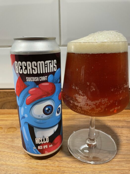 Ölburk med tecknat ansikte bredvid glas med röd IPA-öl och skumtäcke.