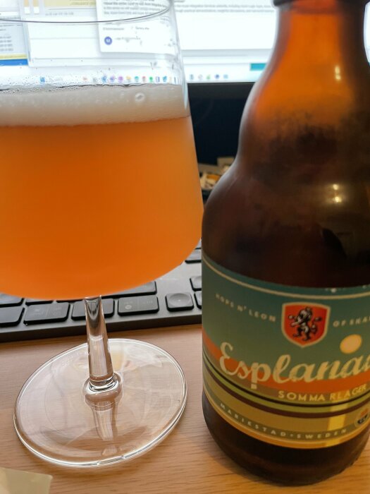 Ölglas med skum, ölflaska, dator och tangentbord i bakgrunden. Inomhus, möjligen arbetsmiljö.