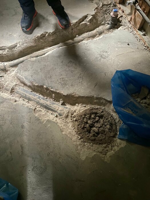 En person står nära grävt hål i betonggolv inne i byggnad under renovering eller reparation.