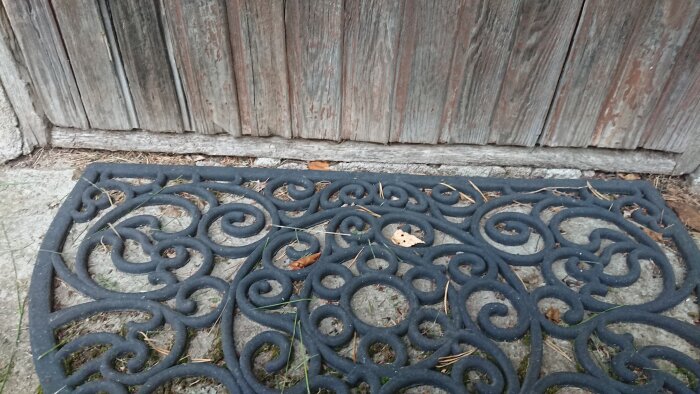 Svart smidesjärnsmatta med spiral-mönster framför en sliten trägrind. Löv och grenar syns på marken.