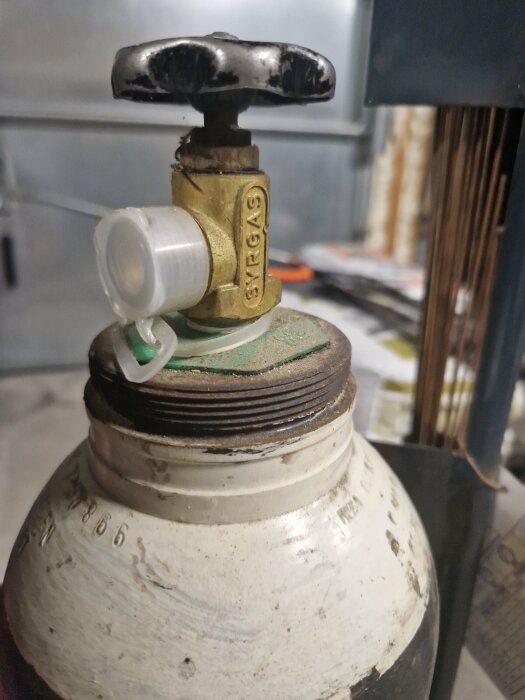 Gammal, använd gasflaska med ventil och märkning SYRGAS. Slitage synligt. Grå, vit och guldtoner dominerar.