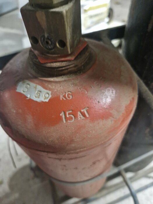 Röd gasflaska med ventil, markerad "5,9 KG" och "15 AT", möjligtvis för gaslagring eller distribution.