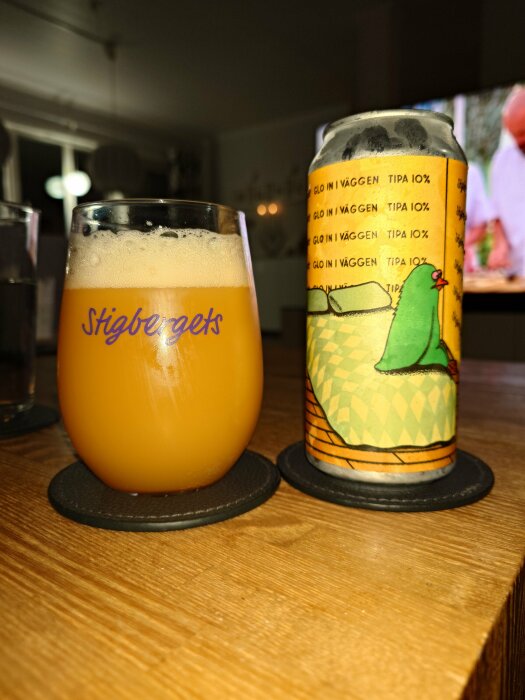 Ölglas med skum topp, burk med färgstark etikett, "Stigbergets" logotyp, inomhus, trämöbel, suddig TV i bakgrunden.