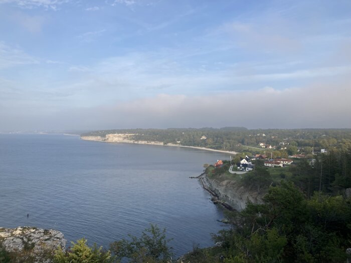 Kustlandskap, klippor, hav, bostadshus, träd, molnigt, lugnt vatten, morgon- eller kvällsljus.
