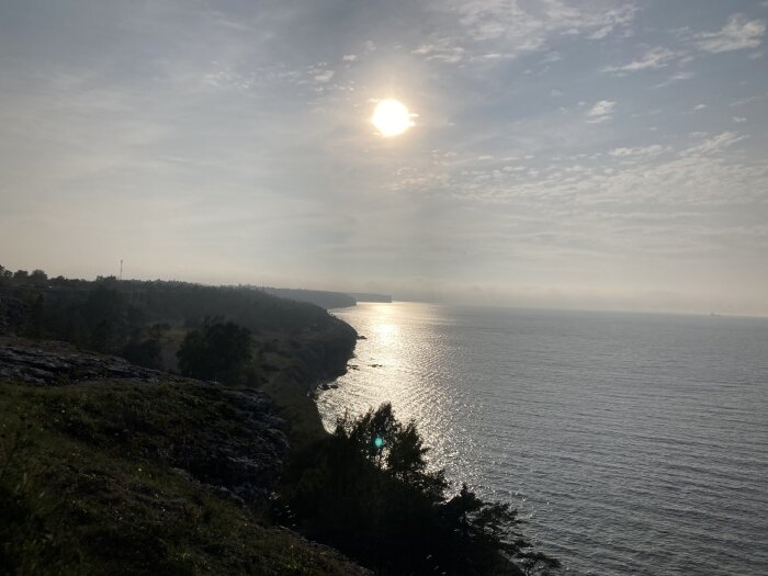 Solnedgång över havet, klippor i förgrunden, moln på himlen, reflektion på vattenytan, lugnt och stilla landskap.