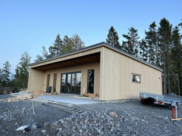 Modernt hus under uppbyggnad med träfasad, stora fönster, skogsbakgrund, släpvagn och byggmaterial på marken.