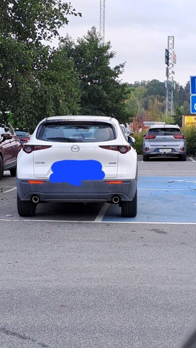 Två vita bilar parkerade, en markerad med handikappskylt. Träd och butiksskyltar i bakgrunden. Registreringsskyltar suddade.