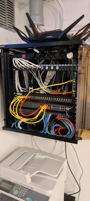 Nätverksrack med switchar, routrar, ethernet-kablar och en wifi-router ovanpå, bredvid ett kopieringsrum.