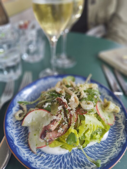 Färgglad salladstallrik med skaldjur, grönsaker, ost; blurrad vitvinsglas i bakgrunden; måltid på restaurang.