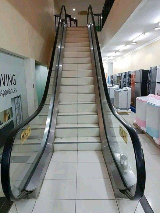 Rulltrappa med trappsteg istället för rörliga delar, inne i en butik med vitvaror. Absurd, ironisk designfel.