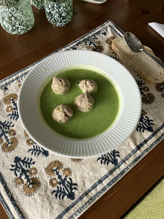 Grön ärtsoppa i vit tallrik med köttbullar, brödskiva, mönstrad duk, glas på bord.