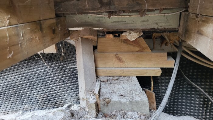 Bild på temporär stödstruktur under ett hus med träbitar och cementblock, sladdar och nät synliga.