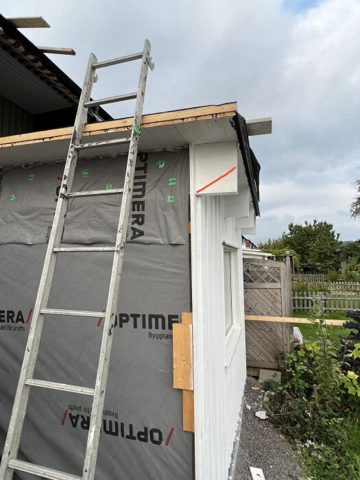 Stege mot en husvägg under renovering, isolering synlig, grå himmel och grönska i bakgrunden.