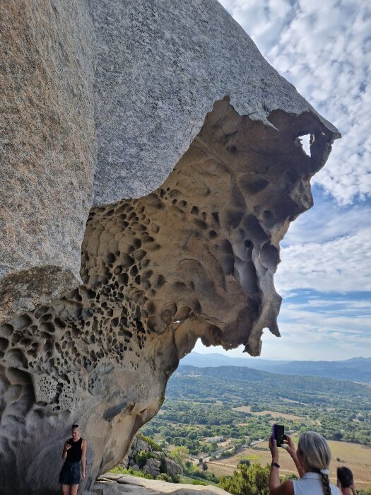 Erosionsformad klippa med hål, personer observerar och fotograferar, vidsträckt landskap i bakgrunden, molnig himmel.