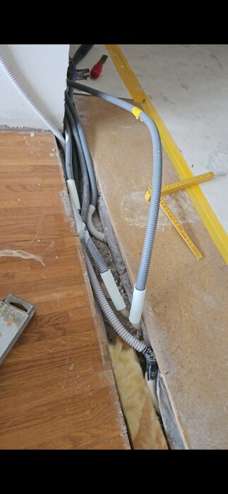 Renoveringsarbete med synliga elkablar och slangar längs en öppen golvkanal vid en vägg.