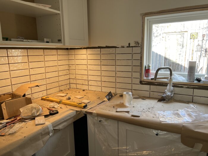 Kök under renovering med kakel på väggen, verktyg och byggmaterial på bänken.