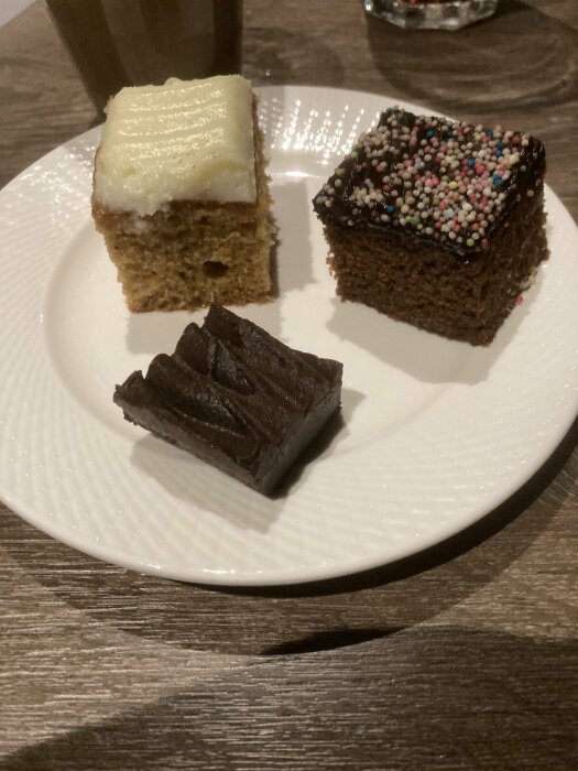 Vit tallrik med tre kakbitar: morotskaka, brownie med strössel och kladdkaka, på ett brunt bord.