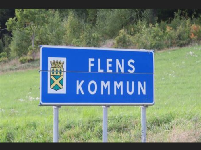 Blått vägskylt med texten "FLENS KOMMUN" och kommunvapen mot grönt landskap.