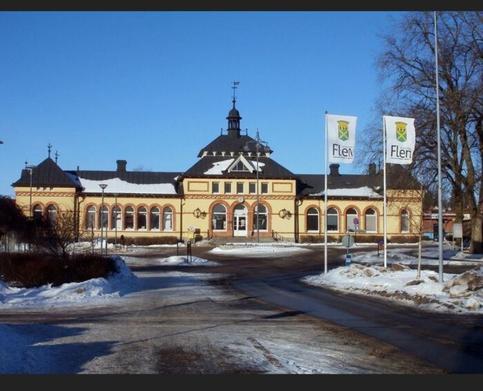 Historisk järnvägsstation, Flen, snökanter, blå himmel, flaggor med kommunvapen, ingen människor synliga, vinterdag.