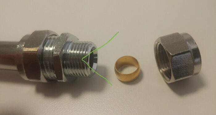 Metallisk rörkoppling med gängade delar och packning, ligger på ett ljusgrått underlag.