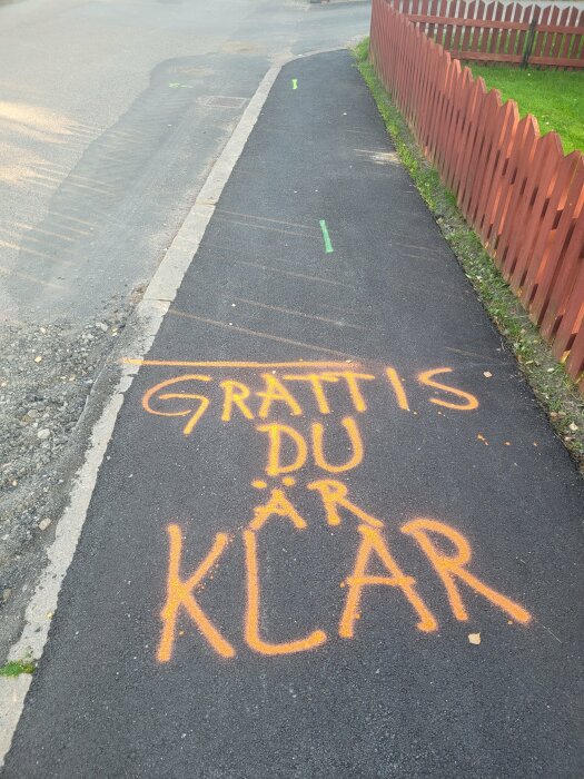 Gatubild med texten "GRATTIS DU ÄR KLAR" målad på trottoaren, staket, grönska, utan människor.