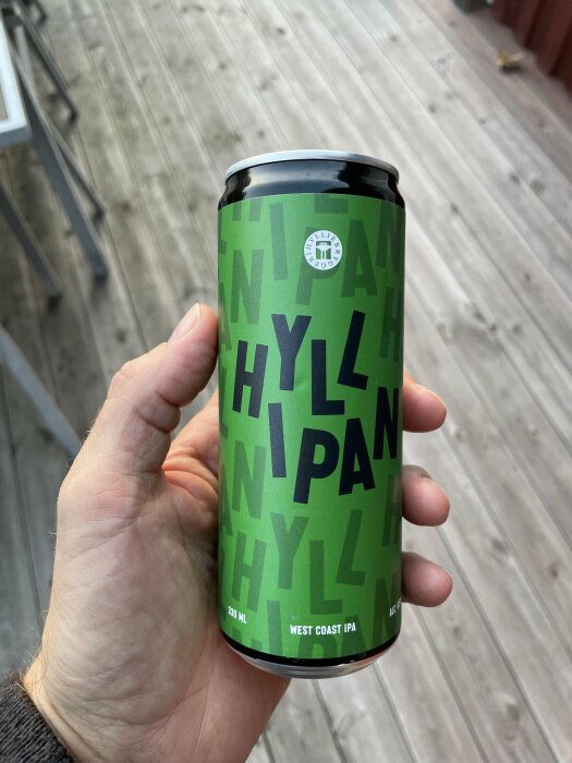 En hand håller en burk West Coast IPA-öl med grön etikett på en träveranda.