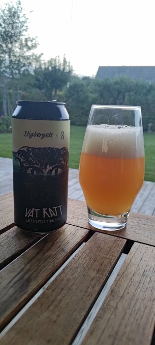 Ölburk och glas på träbord, gräsmatta i bakgrunden, kvällsljus, "Våt Katt", dubbel IPA, skumkrona.