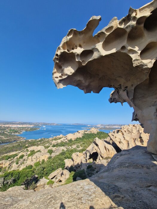 Erosionsformad klippa, klarblå himmel, kustvy, gröna buskar, kustlinje i bakgrunden, klippiga formationer, naturlig arkitektur.