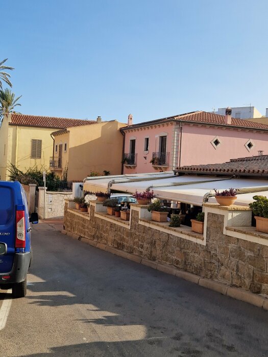 Mediterran gata, rosa och gula hus, palmträd, klar himmel, skuggor på vägen, parkerad blå bil.