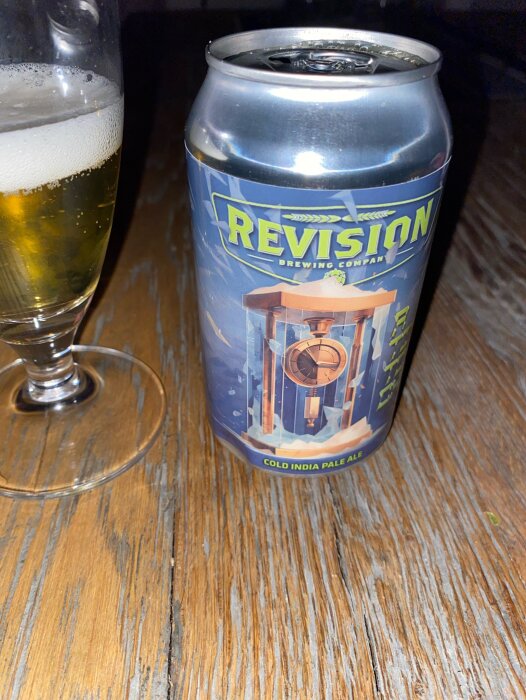 Ölburk med texten "REVISION BREWING COMPANY" bredvid halvfullt ölglas på träbord. Cold India Pale Ale.