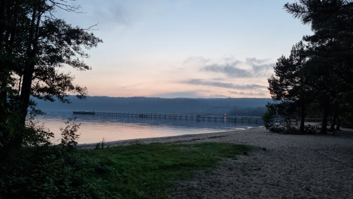 Solnedgång vid sjö med brygga, träd, lugnt vatten, och sandstrand. Tidig kväll, stilla natur, fridfullt landskap.